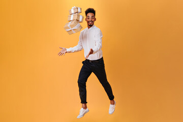 Fototapeta na wymiar Guy in black pants and shirt jumps on orange background with gifts. Full-lenght snapshot of dark-haired man in white shirt on isolated backdrop