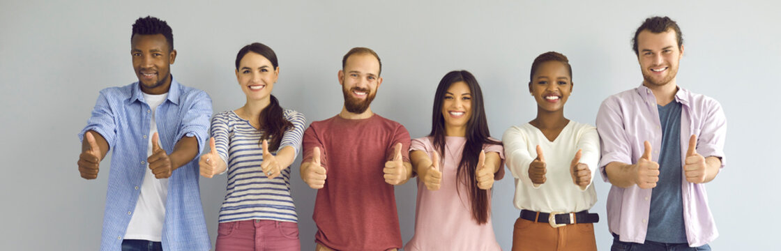 Group portrait of cheerful diverse people giving thumbs up. Team of happy satisfied young men and women standing in row against studio wall and showing like gesture all together. Banner, header image