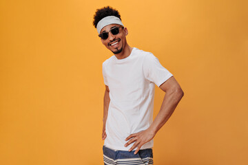 Man in white outfit and dark glasses is smiling on orange background. Portrait of dark-skinned guy in headband and singlasses on isolated backdrop