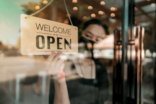 Open Cafe Or Restaurant. Barista Or Waitress Woman Wearing Protective Face Mask And Turning Open Sign Board On Glass Door In Modern Cafe Coffee Shop