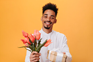 Brown-eyed guy in white shirt holding pink tulips and gift on orange background. Attractive man in...