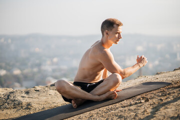 Fototapeta na wymiar Healthy and fit guy with bare muscular torso sitting on yoga mat and stretching body. Handsome sportsman enjoying outdoors workout during morning time.