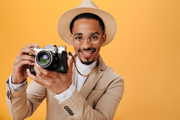 Man in beige hat is holding retro camera on orange background. Cheerful man in jacket and white tee smiling and taking photos on isolated backdrop