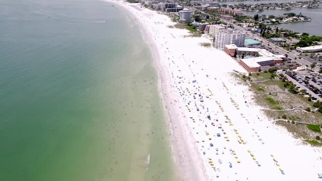 Drone shot of Saint Pete Beach Florida in the summertime. Shot runs along the beach over swimmers showing the town as well as hotels and condos. features green calm water and white sand beach.