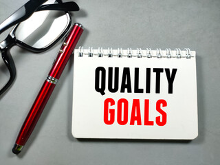 Text QUALITY GOALS on notebook with pen and glasses on gray background.