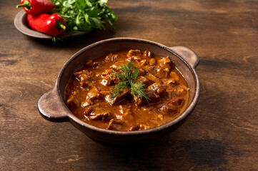 Traditional goulash meat in ceramic bowl on wooden brown background with pepper and herbs. Close up - obrazy, fototapety, plakaty