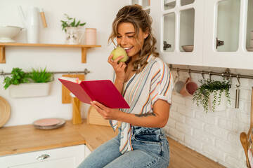 Smiling  blond  woman looking at recipe in cookery book and holding apple in light kitchen at home....