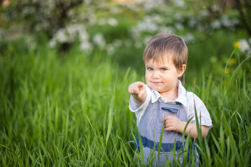 Cute boy in fashionable clothes with blue eyes plays and jumps in the tall grass in a large green blooming garden