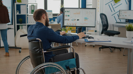 Fototapeta na wymiar Paralized, immobilized, handicapped marketing manager project sitting in wheelchair in start up business office typing on pc checking reports analysing datas. Diverse team discussing financial project