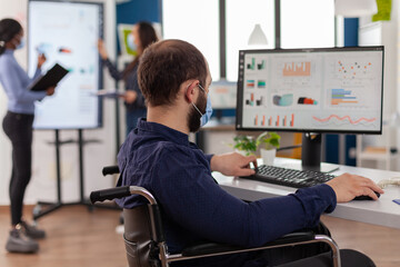 Immobilized business man with protective mask working in new normal business financial company typing on pc, checking reports, analysing data looking at desktop sitting in wheelchair.