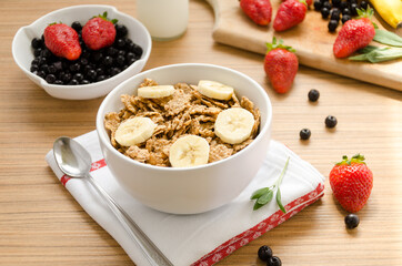 Healthy breakfast with bowl of cereals (muesli), strawberry, wild berries, milk and banana on light wooden table.
