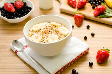 Healthy breakfast with bowl of cereals (muesli), strawberry, wild berries, milk and banana on light wooden table.