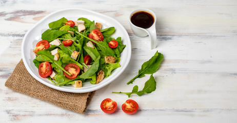 Fresh spinach salad with tomato, small pieces of herb roasted chicken in white ceramic dish on brown sackcloth, together with balsamic vinegar on white wooded table. Concept for healthy