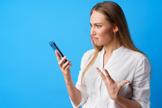 Portrait of young angry woman using her phone, annoyed texting with someone, blue background