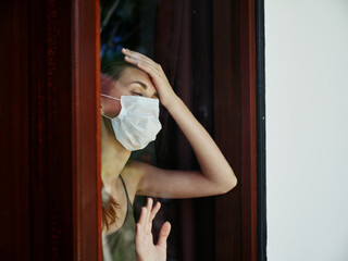 woman in medical mask holds hand on forehead view from window