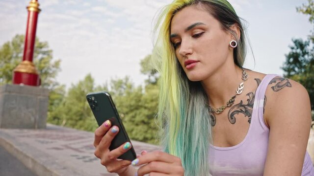 Girl with multicolored yellow blue hair at the park. Close-up portrait of a young woman with bright appearance, tattoos and a tunnel in the ear. Teenager looking at phone screen