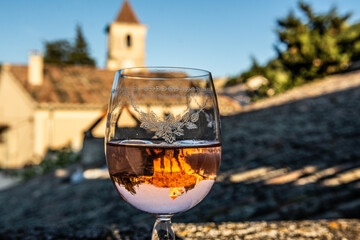 Close up of a glass of rosé wine. View on the roofs of a village in Provence. Church, Roman tiles, blue sky. Inverted reflection of the village, engraved glass.