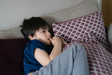 Authentic portrait Kid lying on safa biting his finger nails while watching TV, Little boy lying on...