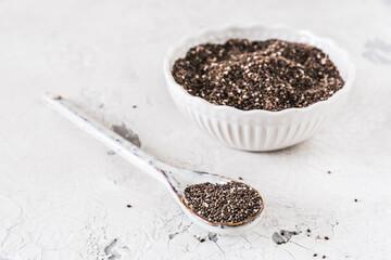 Chia seeds on a white porcellain spoon in front of a white bowl with chia seeds, focus on the spoon, white background, copy space