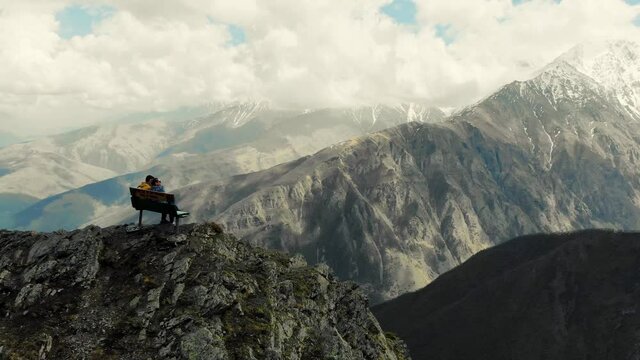 Couple sits on bench at top of mountain, the Caucasus. Bench stands on edge of mountain overlooking huge Caucasus mountains. Snowy peaks of the mountains. Clouds hang from above. Terrible beauty