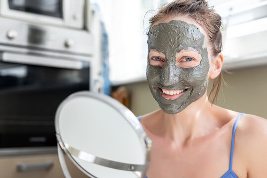 Young adult happy beautiful caucasian woman applying natural gray clay face mask at home against window and looking in mirror. Female person making self facial cleansing moisturizing treatment care