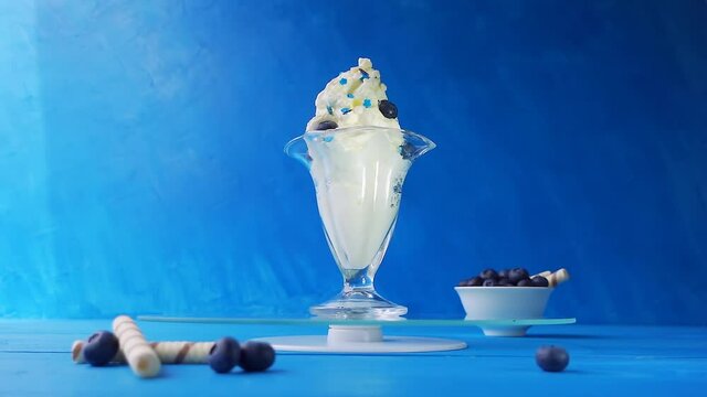Ice cream with blueberries in a beautiful glass glass is spinning on a glass tray on a blue wooden table on a light blue background. Creative slow motion loop video with speed up, down effect.