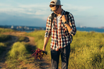 Young handsome man, in a cap and glasses, with a backpack, traveling and walking in nature, a man with a bouquet of wildflowers