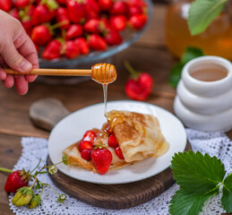 Thin pancakes stacked with strawberries between layers lying on a plate. Hand holding honey spoon and honey flowing on a stack of pancakes