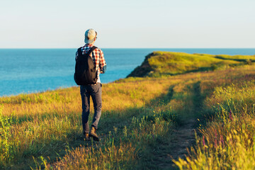 Active tourist, a man with a briefcase in nature, a hipster in a cap walks along a path on a green hill, against the background of the sea