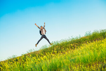 Happy young tourist with a backpack, happy jumping man with hands up against a blue sky background, climbs to the top of a hill or mountain