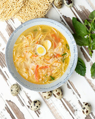 Nutritious soup with noodles and quail eggs on rustic table. Clay bowl of delicious food on white wooden background. Flat lay or top view.