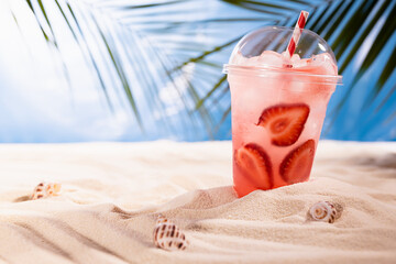 Fresh strawberry beverage with ice, straw in plastic glass on sandy tropical beach with sea view, palm leaves, copy space. Vacation and relax on coast.