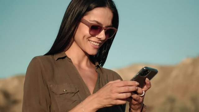 A smiling woman wearing sunglasses is using her mobile while walking outdoors at summer