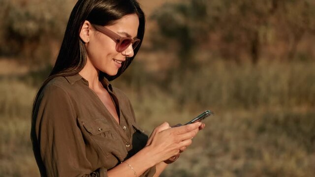 A side view of a smiling woman wearing sunglasses is using her mobile while standing outdoors at summer