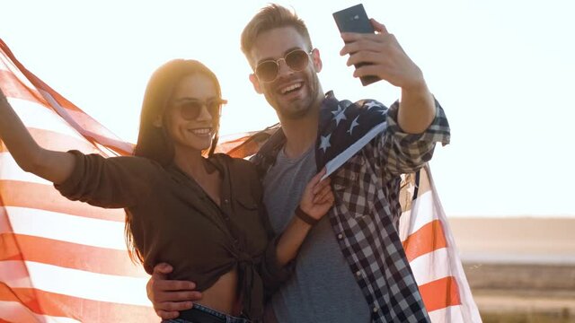 A happy couple man and woman are taking selfie photo with an american flag standing outdoors at summer