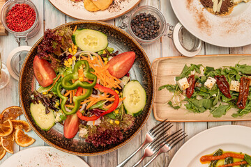 Top view image of vegetable salad and raw fresh vegetables, arugula salad with feta cheese and dried tomatoes, forks, red pepper, black pepper, orange dry