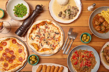 Set of pasta dishes and Spanish food on wooden table. Croquettes, peas with ham, beef meatloaf in sauce, pepperoni pizza, sautéed noodles. Chicken stewed with rice.