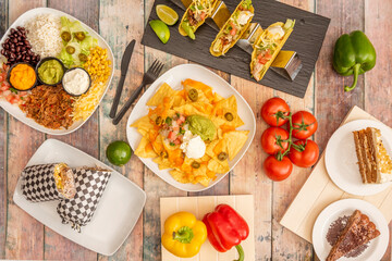 Set of colorful Mexican dishes with corn nachos, avocado guacamole, sweet corn, cheddar cheese, pico de gallo, crispy corn tacos, string tomatoes, full burrito, tricolor peppers on wooden table.