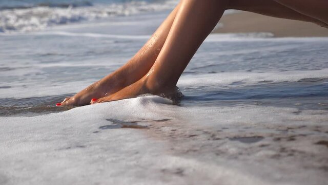 Ocean waves washing over tanned female feet. Legs of sexy girl lying golden sand on sea beach and relaxing during summer vacation travel. Trip and relax concept. Slow motion Close up