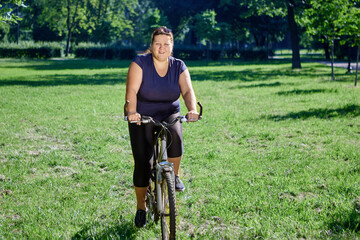 Obese woman in body positivity style rides bicycle on lawn on sunny summer day.
