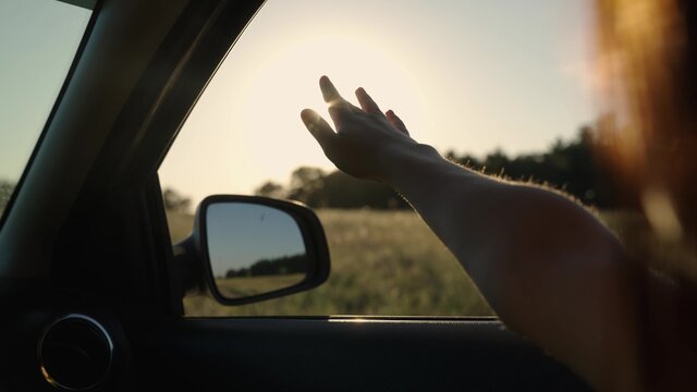 Girl with long hair is sitting in front seat of car, stretching her arm out window and catching glare of setting sun. Free woman travels by car catches wind with her hand from car window. Vacation