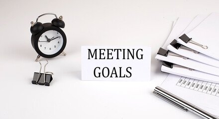 Card with text MEETING GOALS on a white background, near office supplies and alarm clock. Business concept.