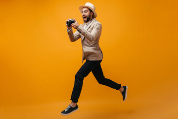 Man in beige jacket and hat jumps on orange background and holds camera