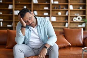 Frustrated indian man feel doubts and despair, sits on the sofa with upset face expression and...