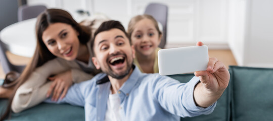 Obraz premium selective focus of smartphone in hand of cheerful man taking selfie with happy wife and daughter, banner
