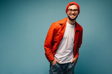 Cheerful man in glasses, hat and orange jacket smiles in blue background