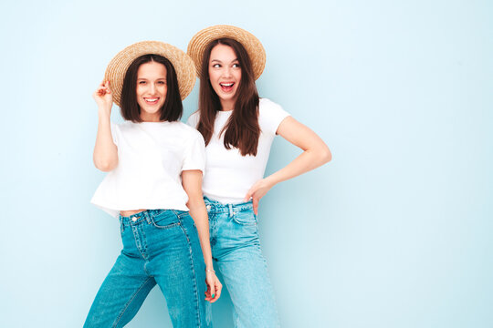 Two young beautiful smiling hipster female in trendy summer white t-shirt and jeans clothes.Sexy carefree women posing near light blue wall in studio.Positive models in hats. Happy and cheerful