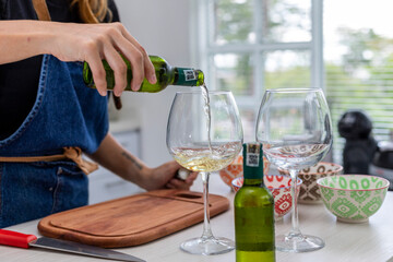 woman in the kitchen pouring two glasses of white wine while she cooks