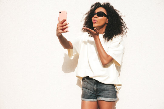Beautiful black woman with afro curls hairstyle.Smiling hipster model in white t-shirt. Sexy carefree female posing in the street near white wall in sunglasses. Cheerful and happy.Taking selfie photo