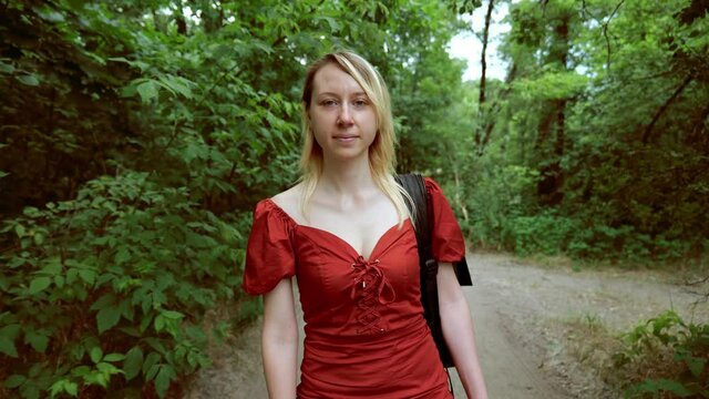Portrait of a blonde woman in a red dress is walking along the road in the forest
